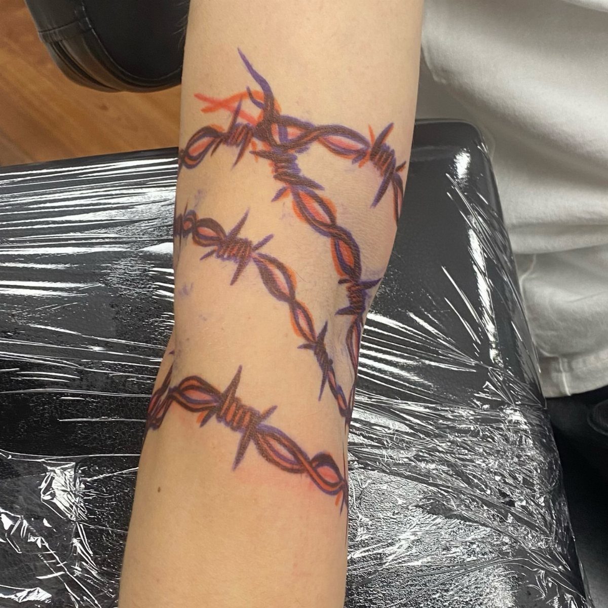 7 Arm Band Colored Tattoo Designs for Men in 2026 6 A forearm displaying a colorful barbed wire tattoo design in red and purple ink.