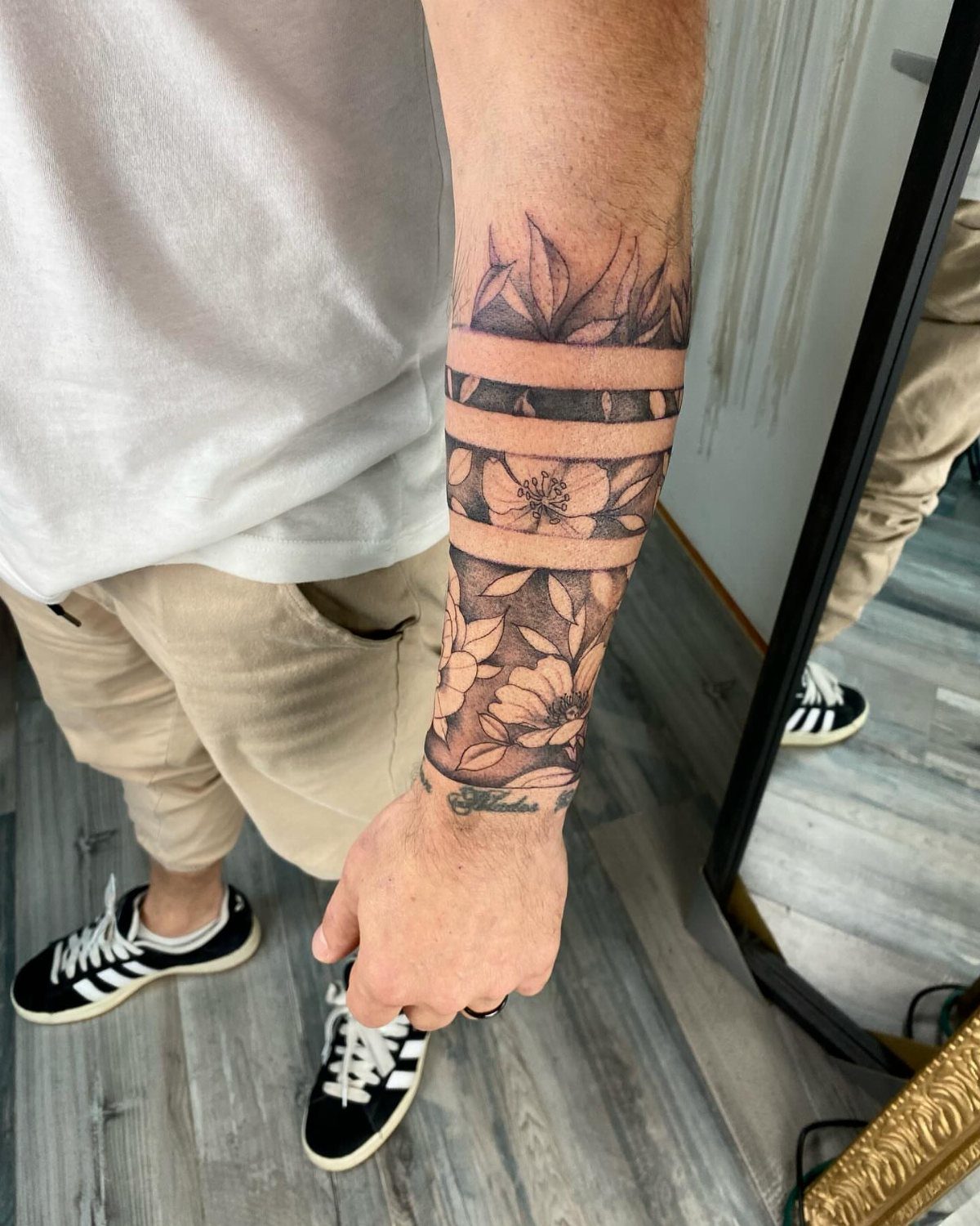 7 Arm Band Colored Tattoo Designs for Men in 2026 2 A man's forearm displaying a black and grey floral armband tattoo with negative space bands.