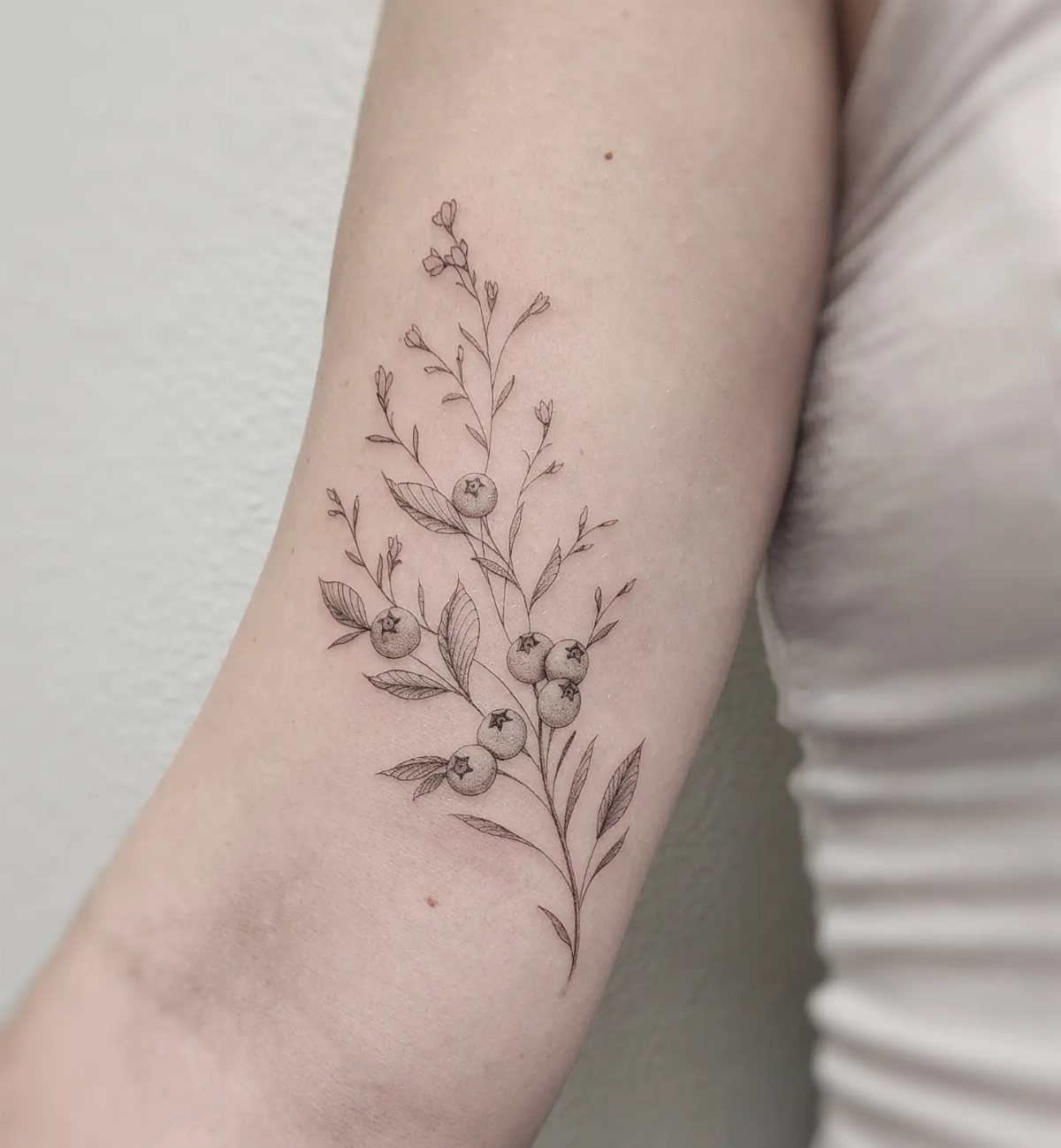 8 Blueberry Tattoo Ideas for Arms, Collarbones and Small Spaces 3 A fine line blueberry branch tattoo with leaves and berries on a person's forearm.