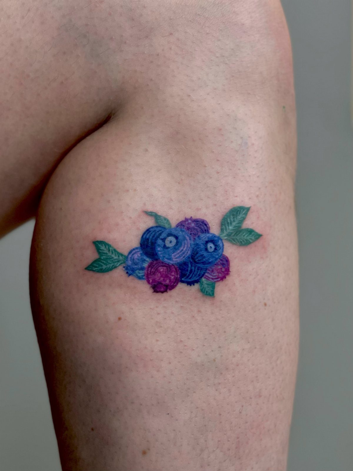 8 Blueberry Tattoo Ideas for Arms, Collarbones and Small Spaces 2 A colorful blueberry cluster tattoo with green leaves on a person's skin.