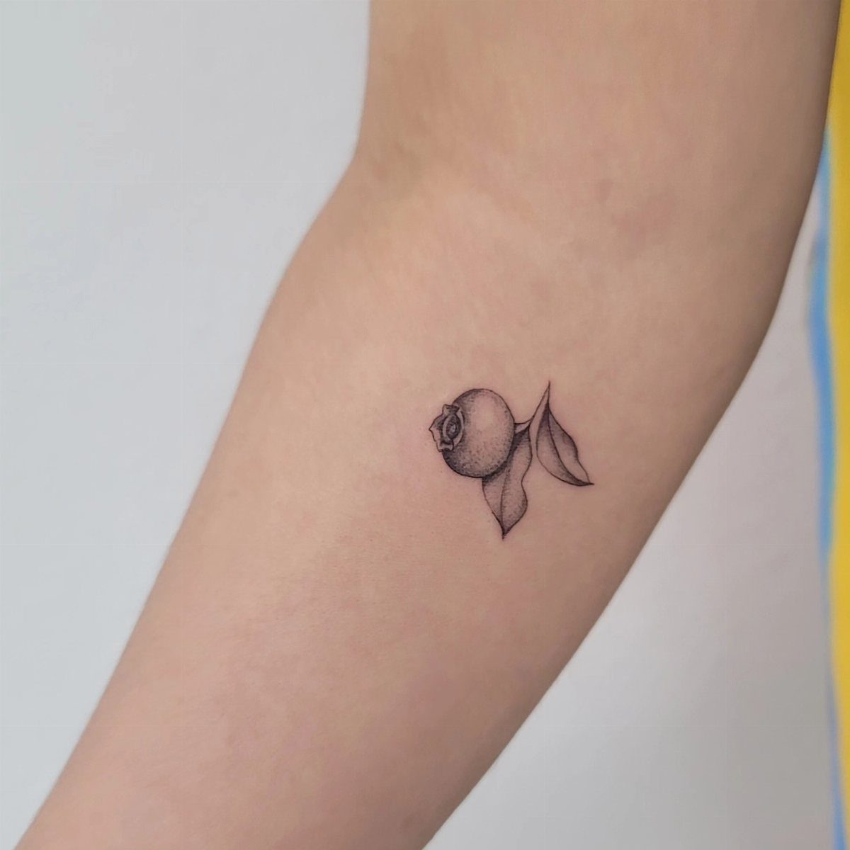 8 Blueberry Tattoo Ideas for Arms, Collarbones and Small Spaces 6 A small fine line tattoo of a single blueberry with leaves on skin.