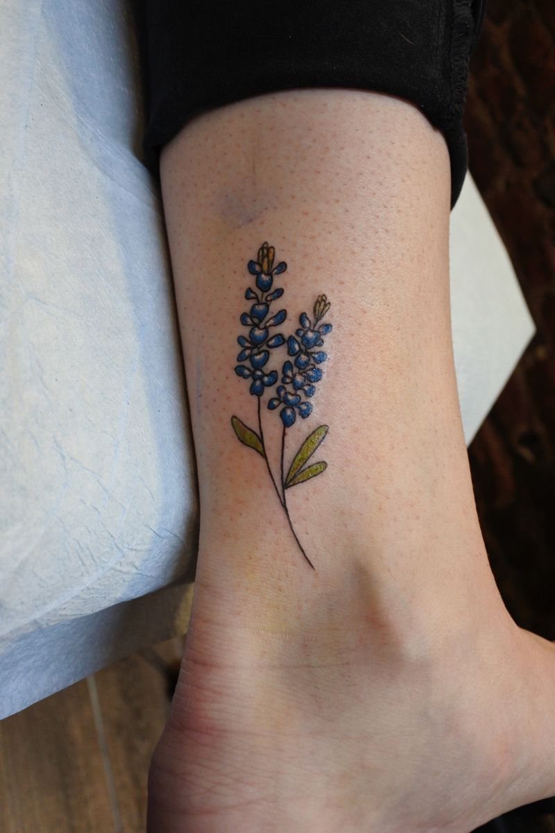A bluebonnet flower tattoo with blue blossoms and green leaves on a person's ankle.