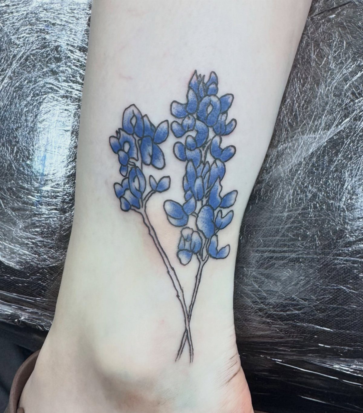 Bluebonnet flower tattoo with two blue stalks on a person's lower leg skin.