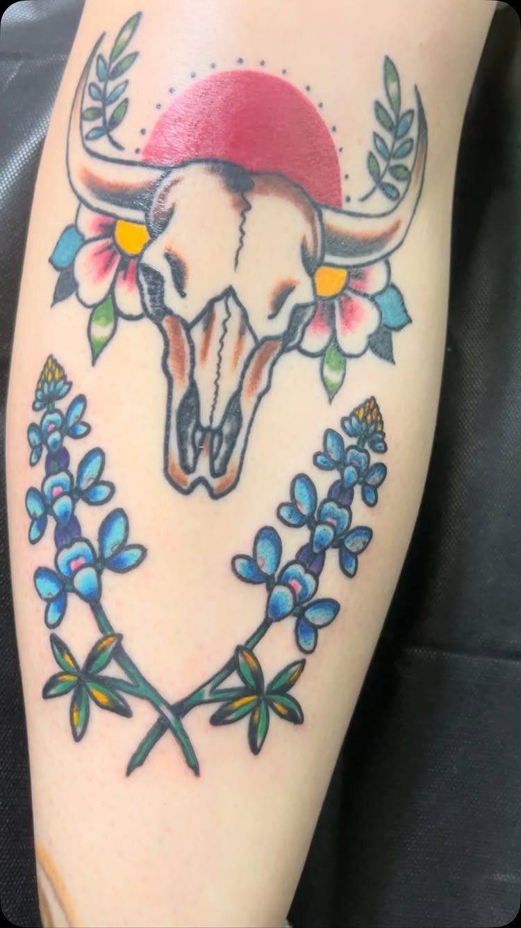 Traditional style tattoo of a steer skull, pink flowers, and bluebonnet stems on a person's leg.
