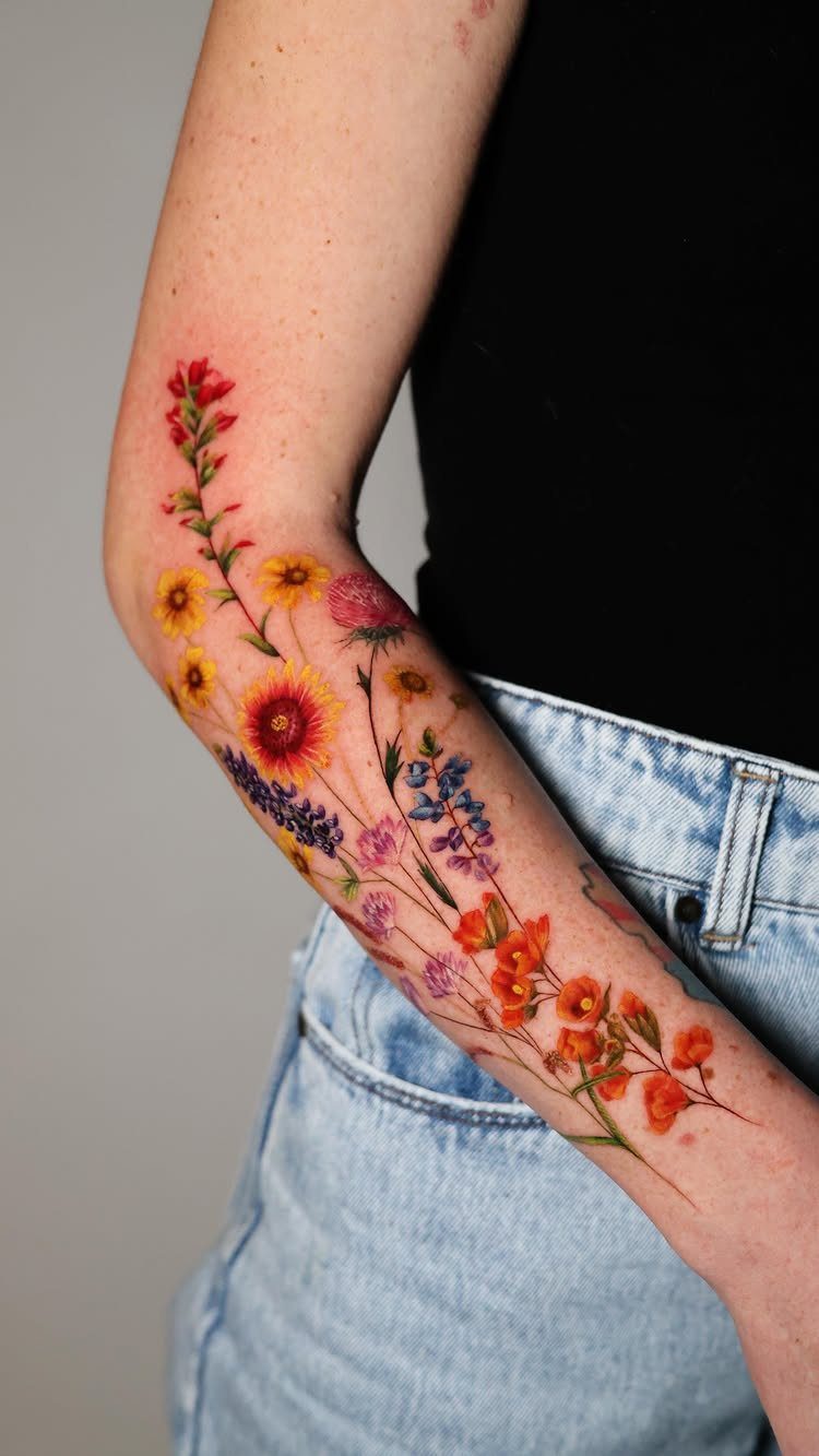 A colorful wildflower tattoo featuring red, yellow, and blue flowers stretching down a person's forearm.