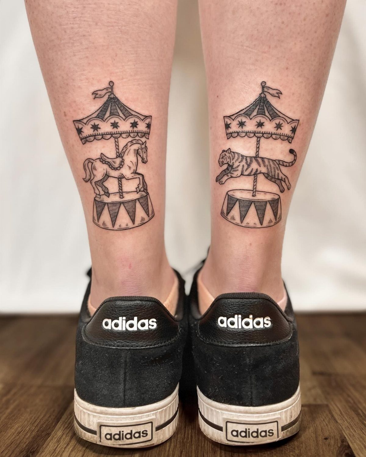 Two black ink minimalist carousel tattoos on a person's lower legs above black sneakers.
