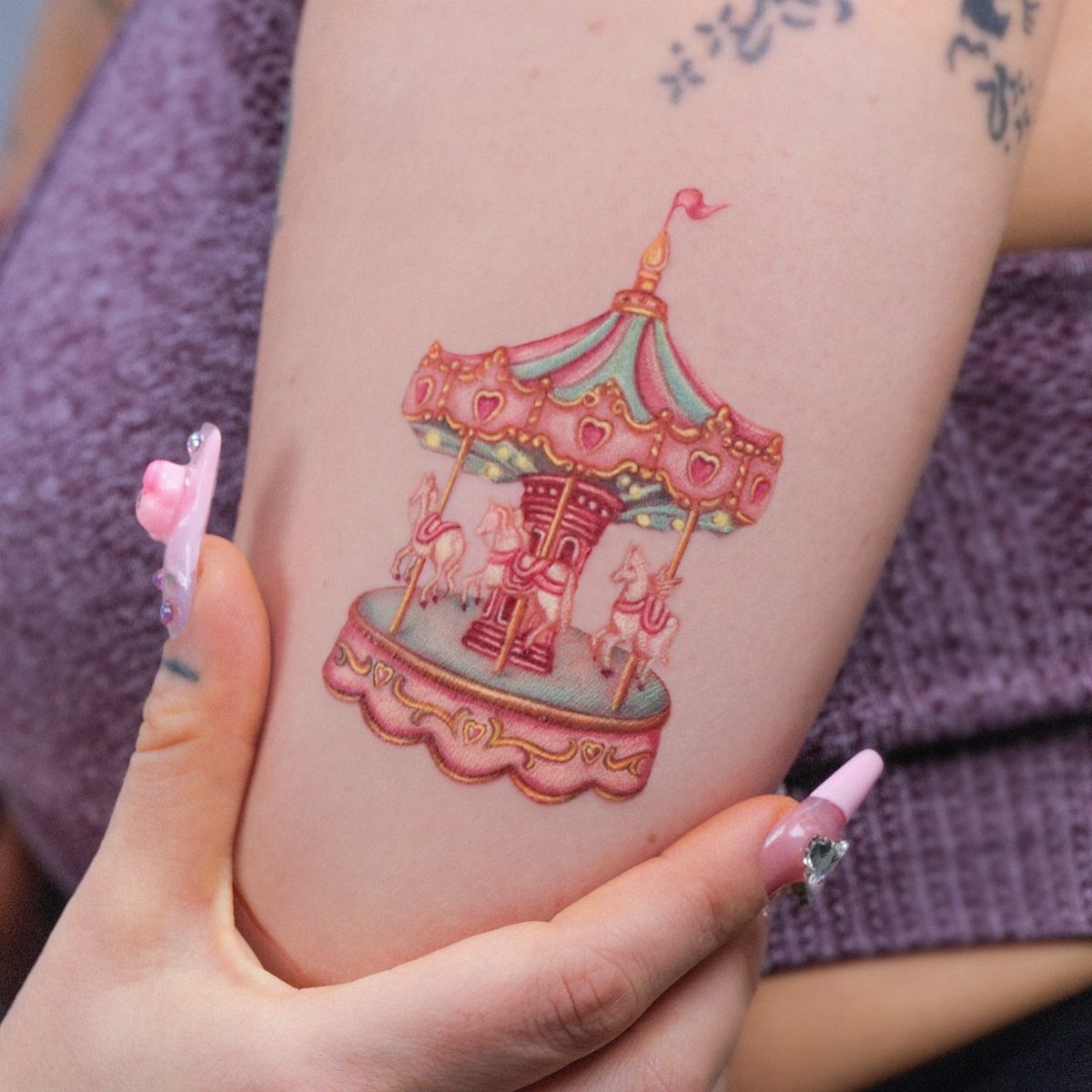 A colorful carousel tattoo with pink and teal accents featuring small horses and heart shapes on skin.