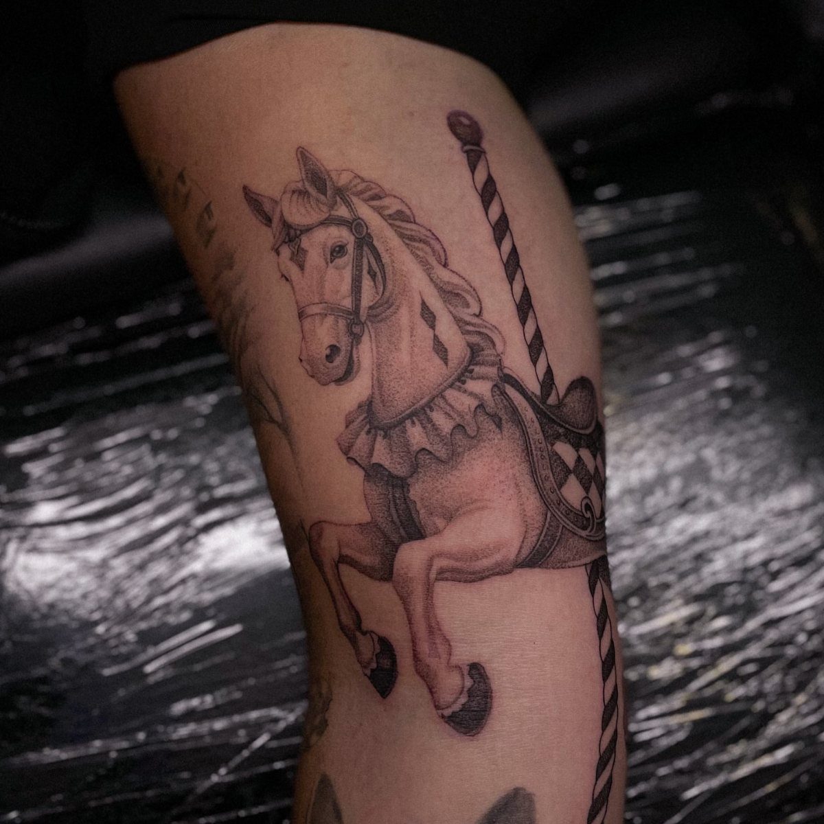 A black and grey tattoo of a single carousel horse on a person's leg.