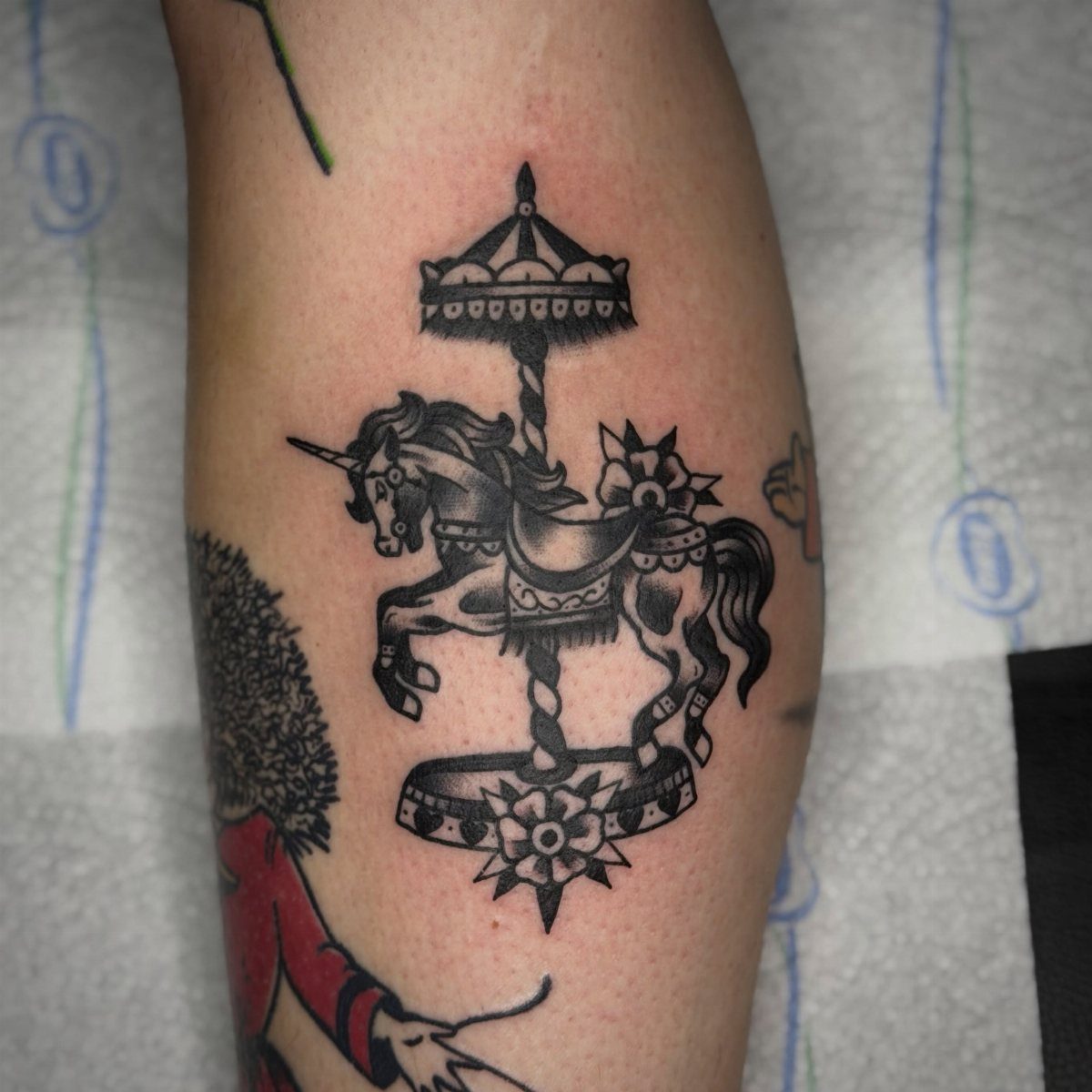 A black ink tattoo of a carousel featuring a decorated unicorn on a person's arm.
