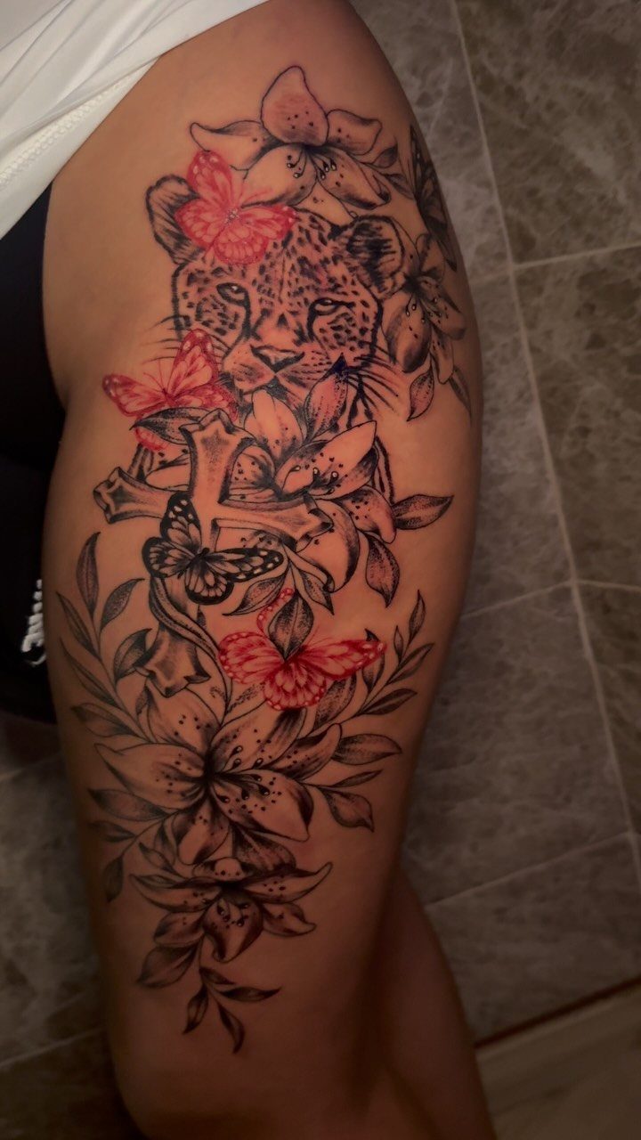 Cheetah Thigh Tattoo: 6 Ideas for Women Who Want Bold Ink 4 A large cheetah head tattoo on a thigh surrounded by lilies and red butterflies.