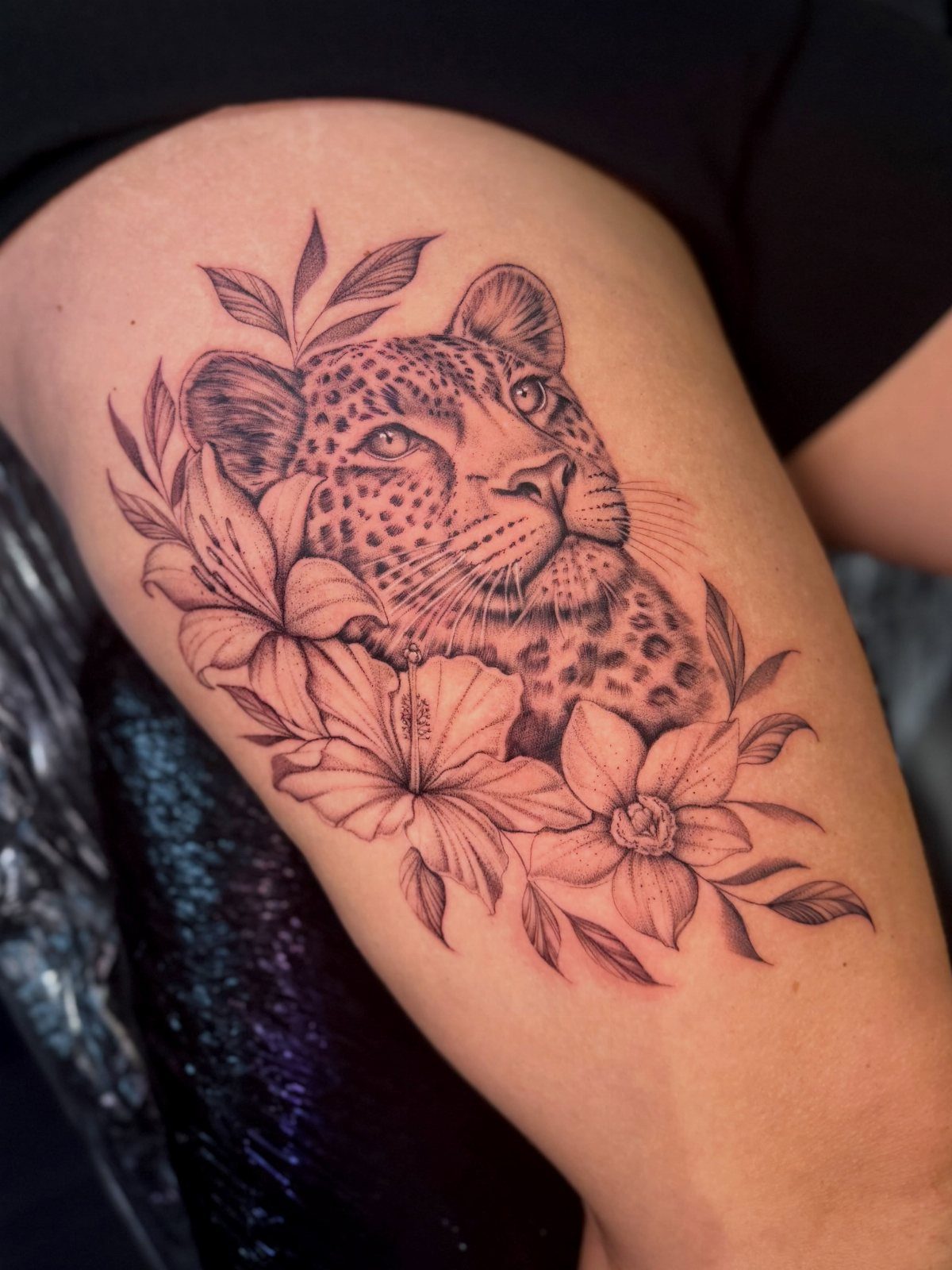 Cheetah Thigh Tattoo: 6 Ideas for Women Who Want Bold Ink 3 A detailed black and grey cheetah head tattoo surrounded by hibiscus flowers on a person's thigh.