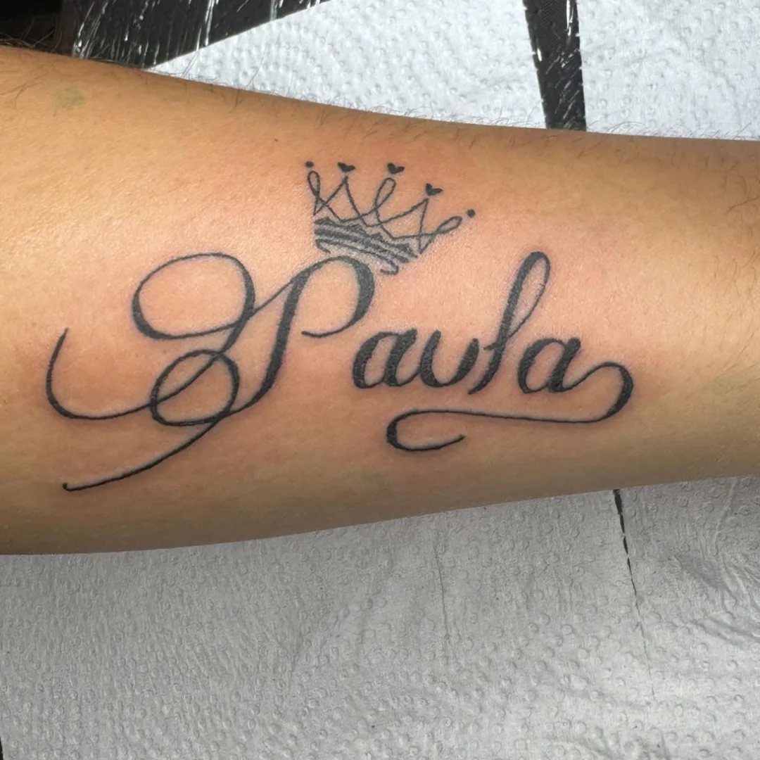 A cursive tattoo of the name Paula with a small crown on a person's arm.