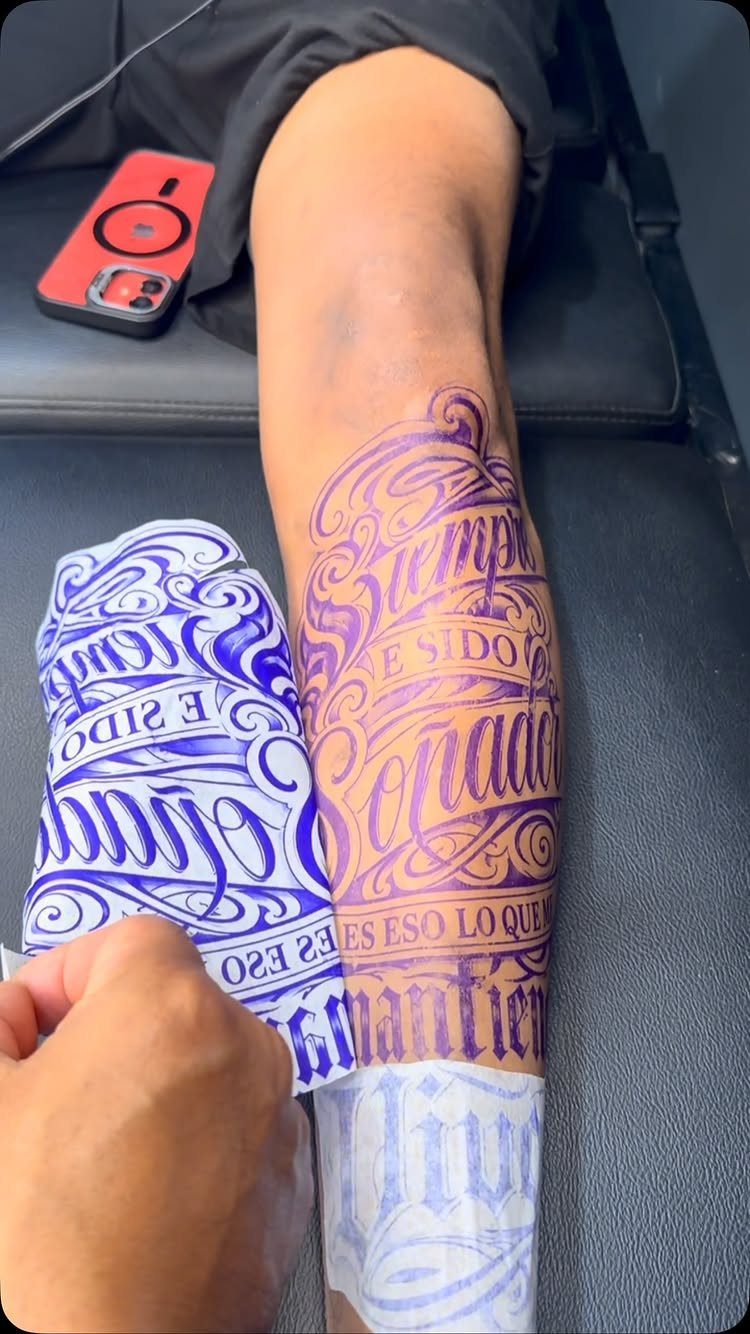 Intricate purple cursive lettering tattoo being applied to a person's leg next to a paper stencil.