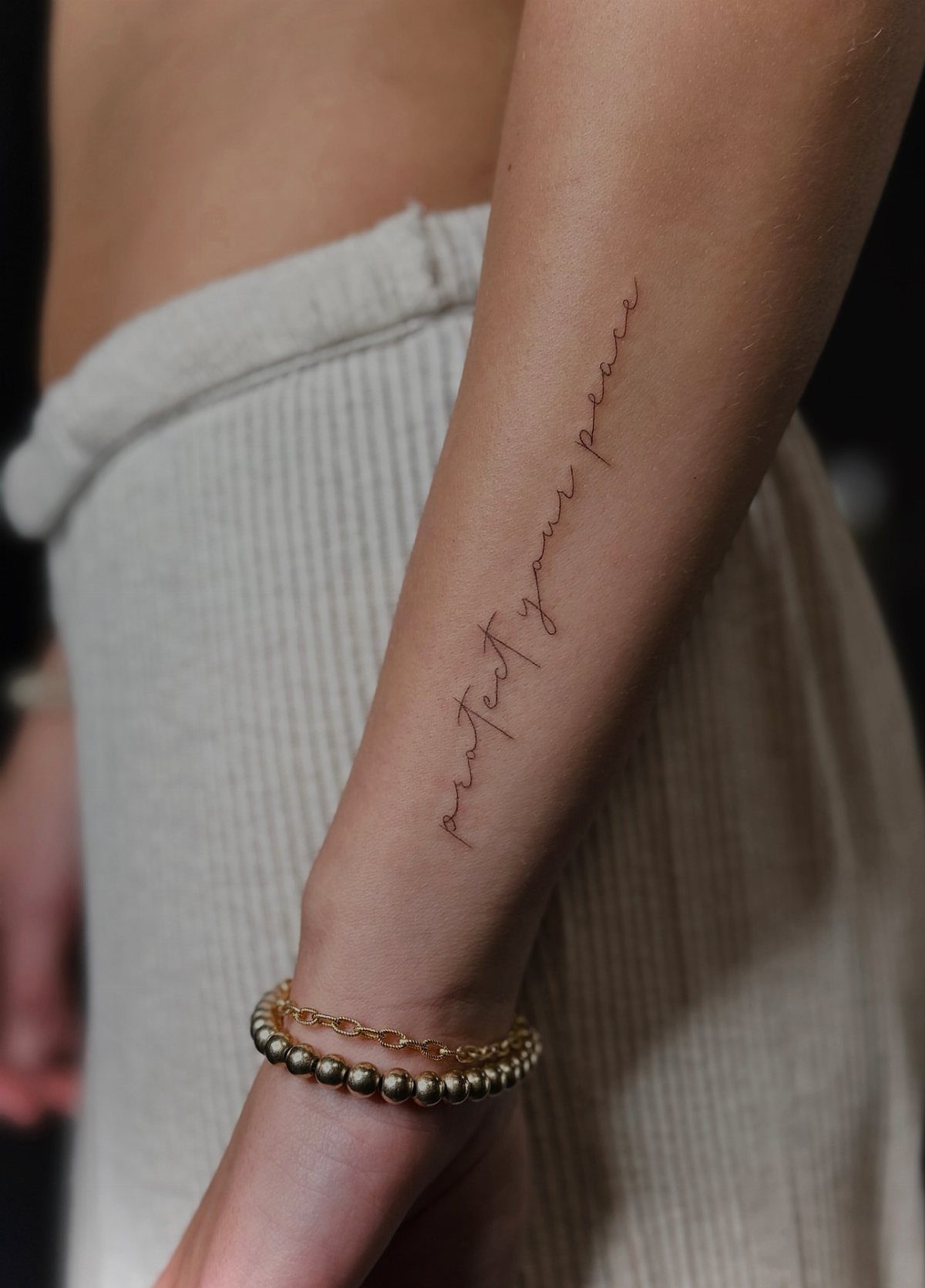 A fine-line cursive tattoo reading protect your peace on a person's forearm.