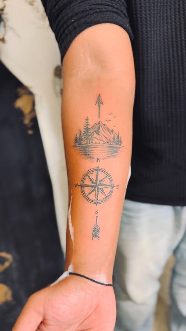 9 Earth Element Tattoo Ideas for Symbols, Scenery and Spirit 8 A black ink tattoo on a forearm featuring mountains, pine trees, and a compass rose.