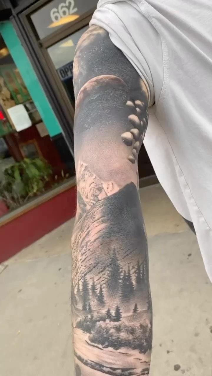 9 Earth Element Tattoo Ideas for Symbols, Scenery and Spirit 6 A detailed black and grey landscape tattoo featuring mountains, pine trees, and a moon on an arm.