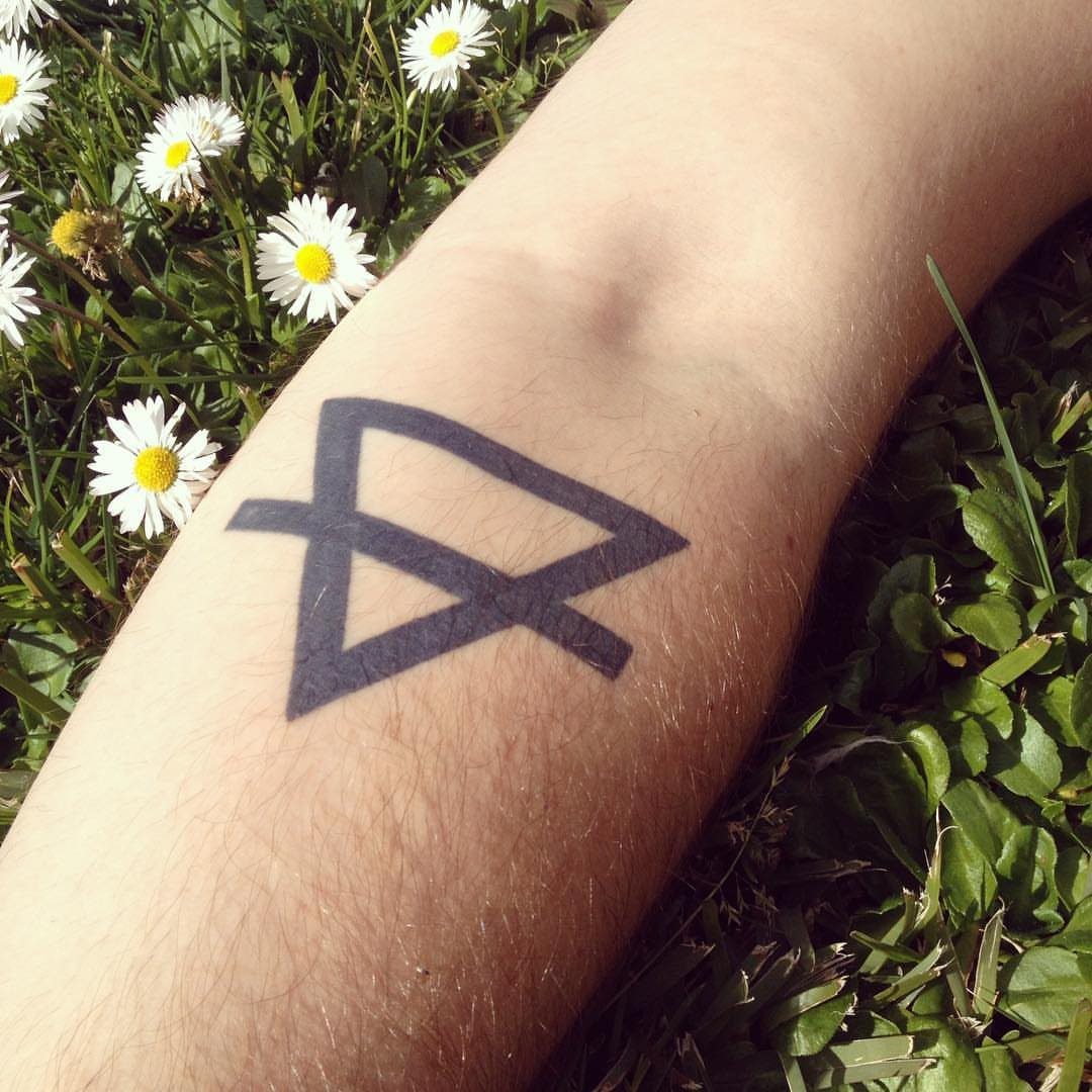 11 Elemental Tattoo Ideas for Your Next Ink 4 A minimalist black geometric symbol tattoo on a person's arm amidst green grass and daisies.