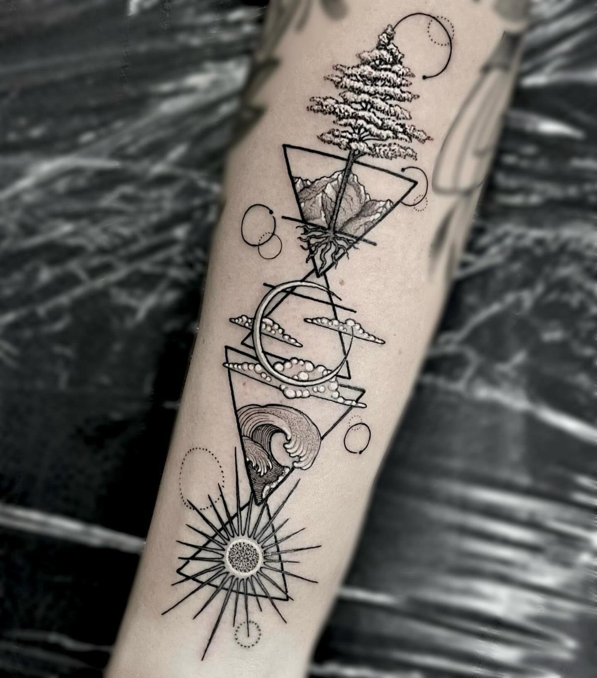 11 Elemental Tattoo Ideas for Your Next Ink 8 A black ink geometric tattoo on an arm featuring trees, mountains, waves, and a sun.