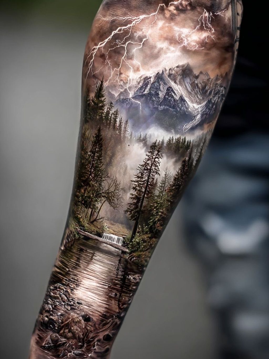 11 Elemental Tattoo Ideas for Your Next Ink 2 A detailed landscape tattoo on an arm showing mountains, forests, a river, and lightning.