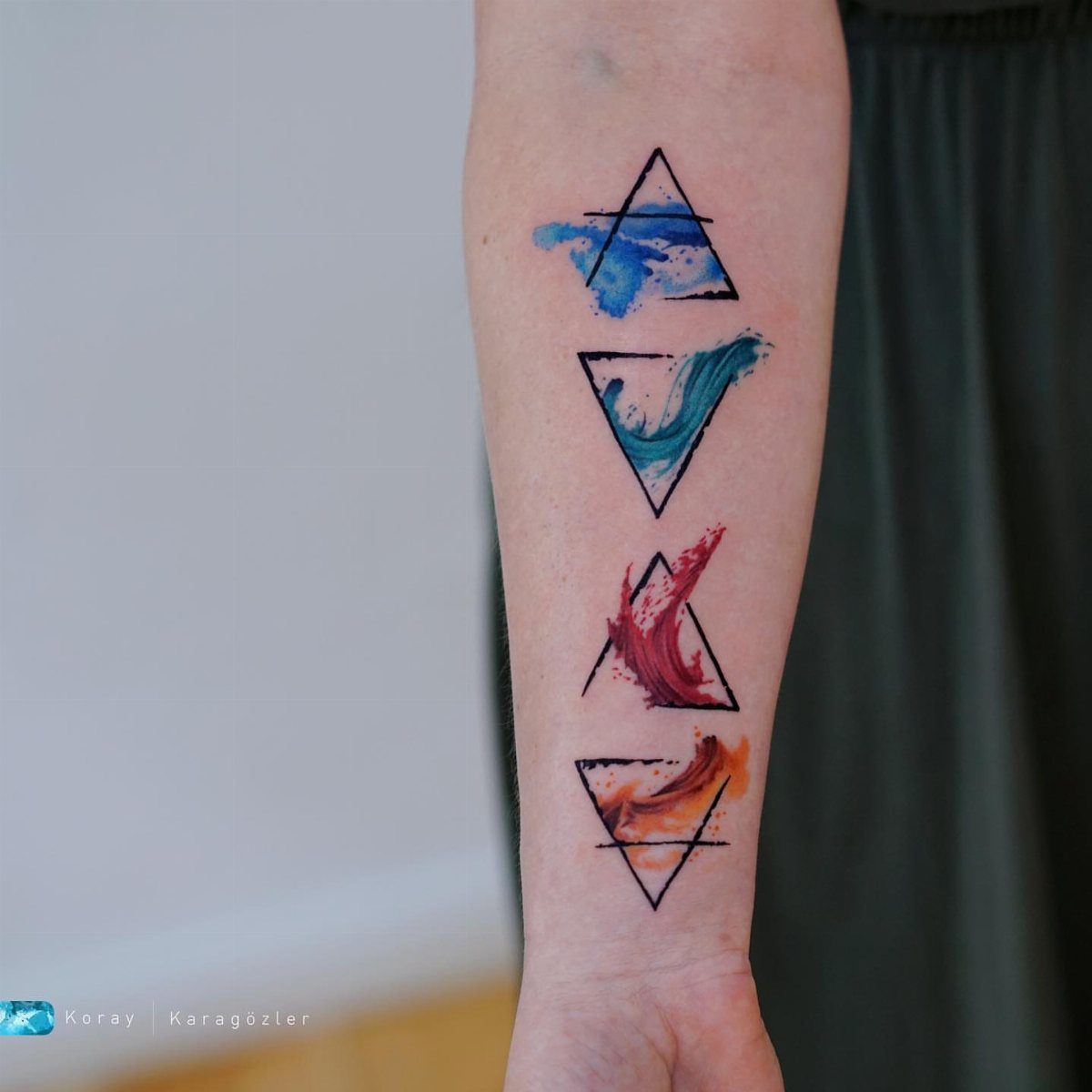 11 Elemental Tattoo Ideas for Your Next Ink 6 Four elemental symbol tattoos in black triangles with colorful watercolor splashes on a person's forearm.