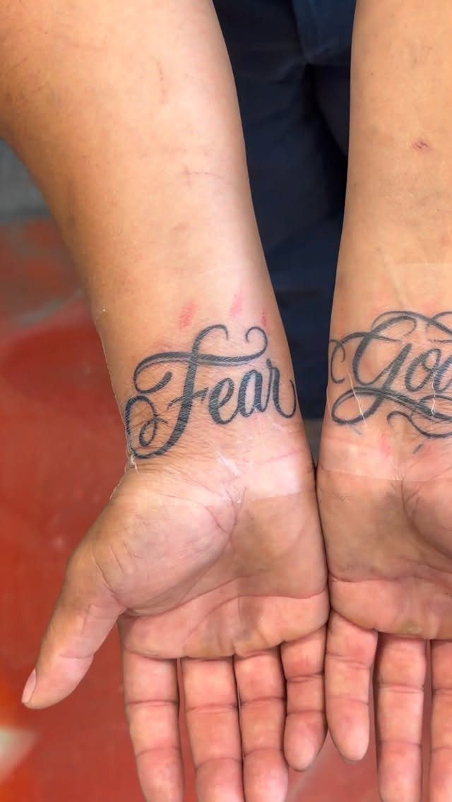 Two wrists showing black ink cursive tattoos of the words Fear God.