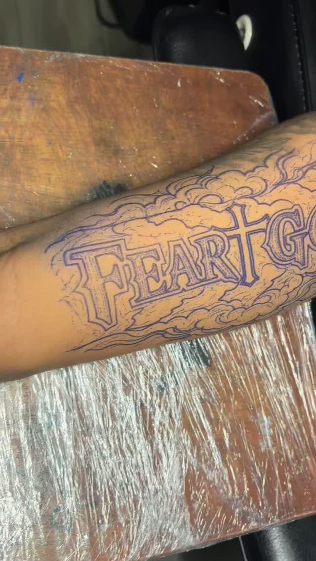 Blue ink tattoo stencil of the words Fear God surrounded by clouds on a forearm.