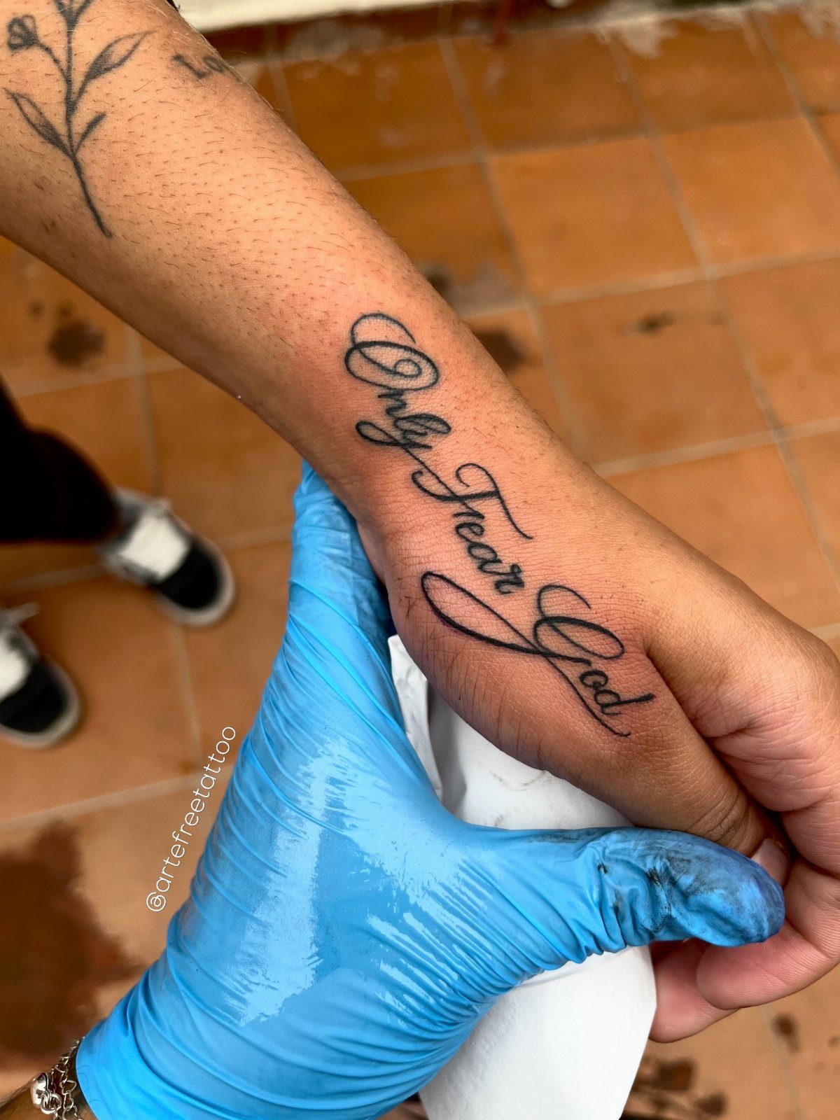 A cursive script tattoo reading 'Only Fear God' on a person's wrist held by a gloved hand.
