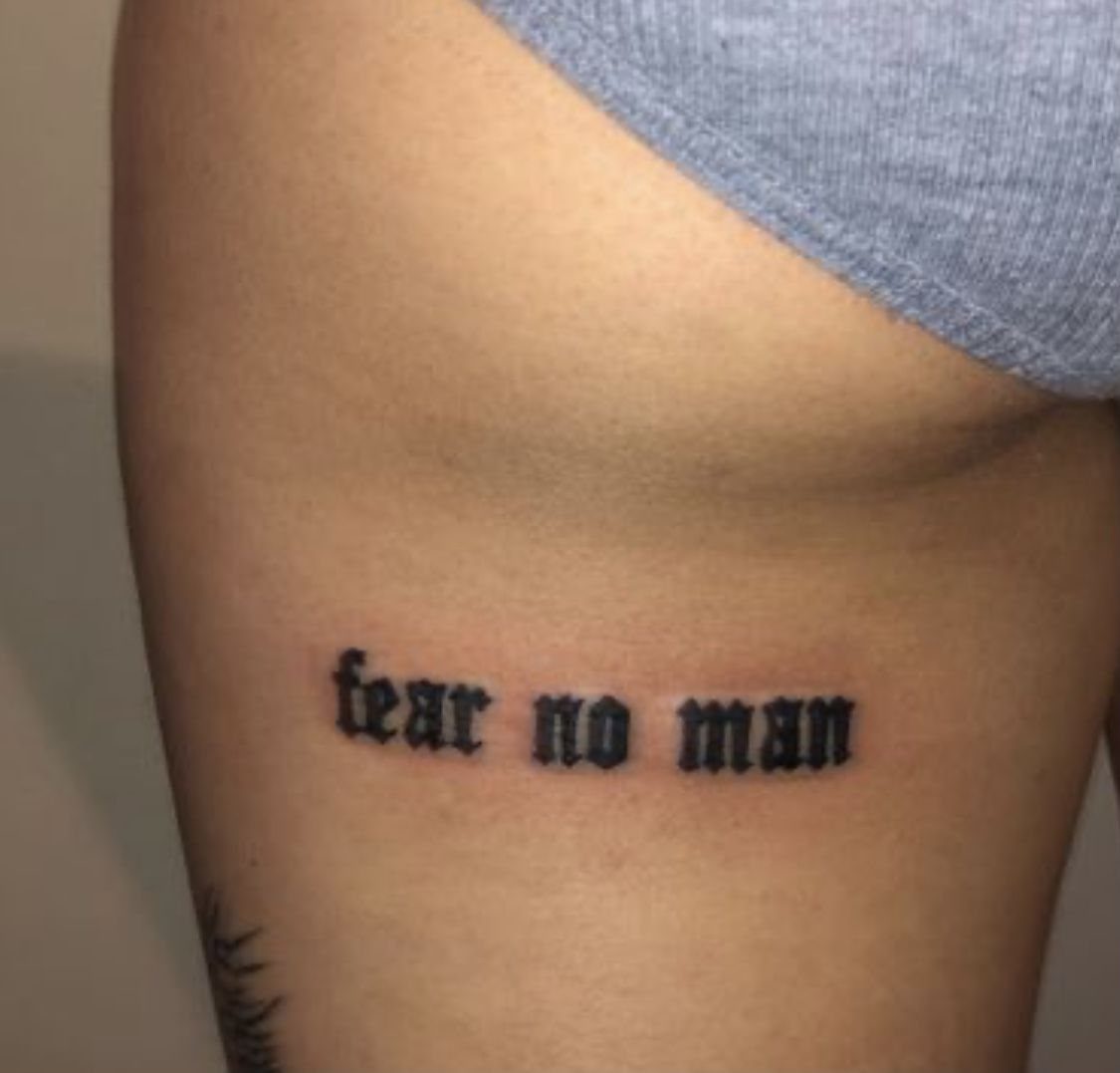 A black gothic font tattoo reading fear no man on a person's hip skin.