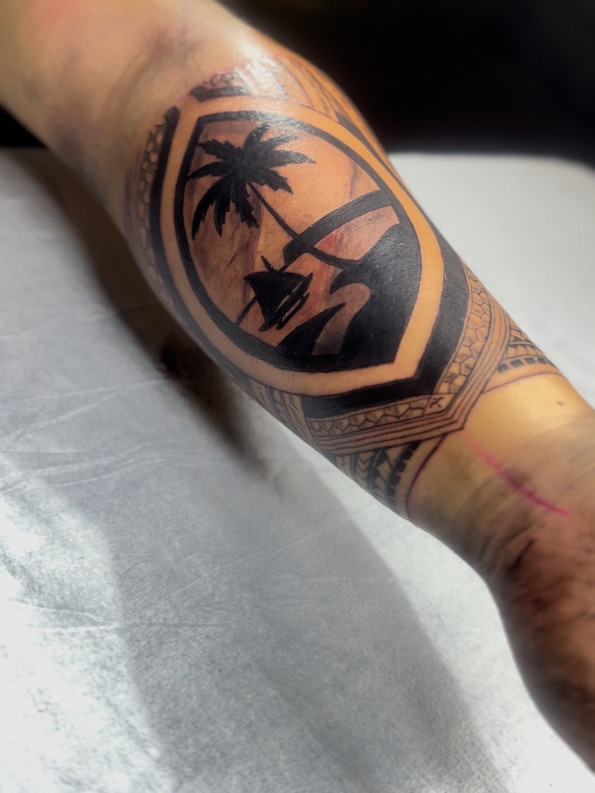 A black ink forearm tattoo depicting a tropical landscape with a palm tree and boat.
