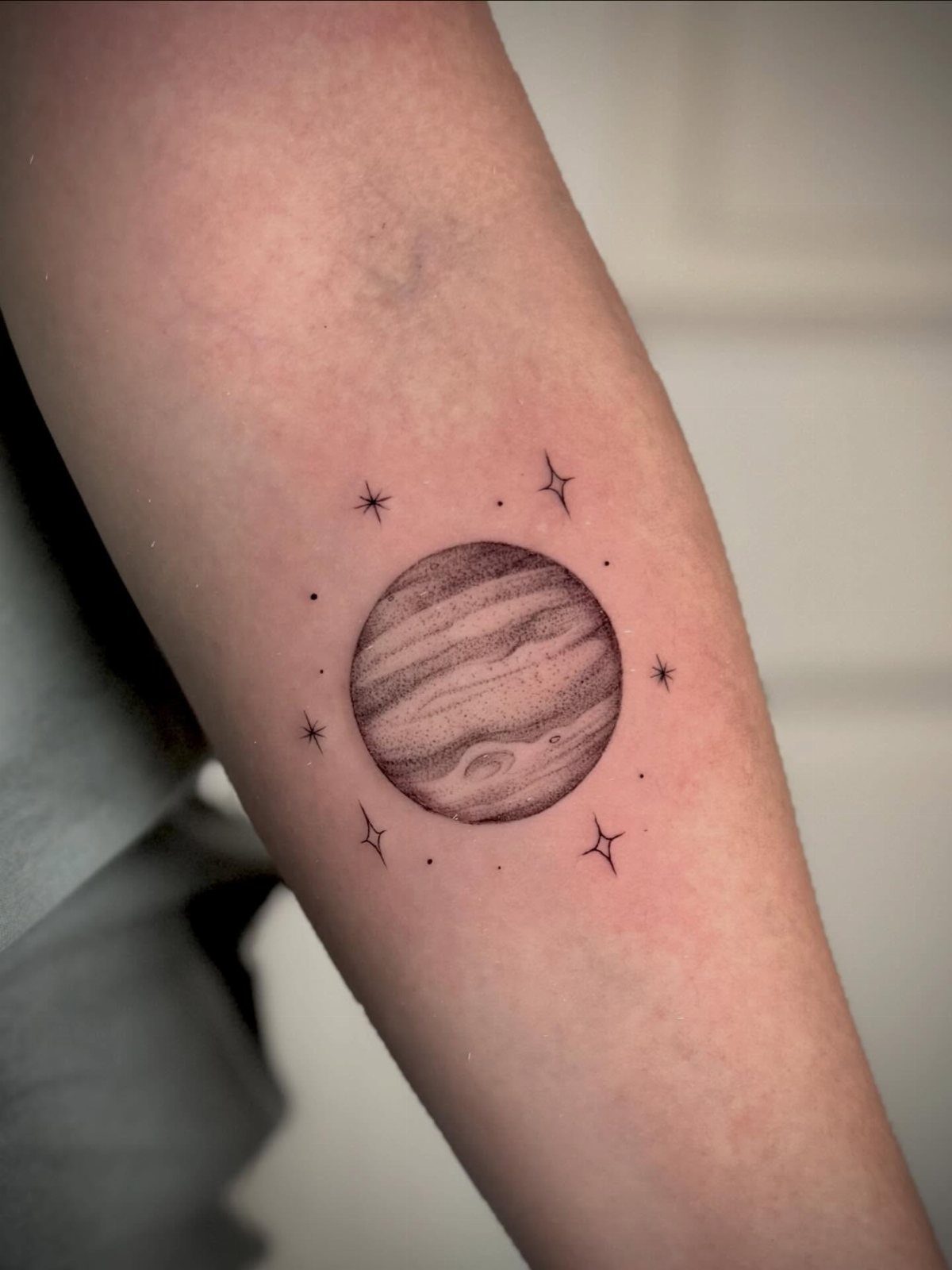 5 Jupiter Tattoo Ideas for Celestial Ink That Stands Out 1 A fine-line black ink tattoo of Jupiter with stars on a person's arm.