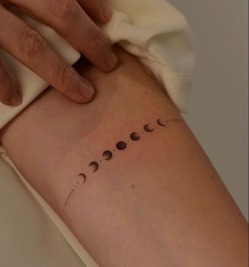 Moon Phases Tattoo Designs for Women: 8 Ideas to Consider 2 A horizontal line of small black moon phase tattoos on a person's arm.