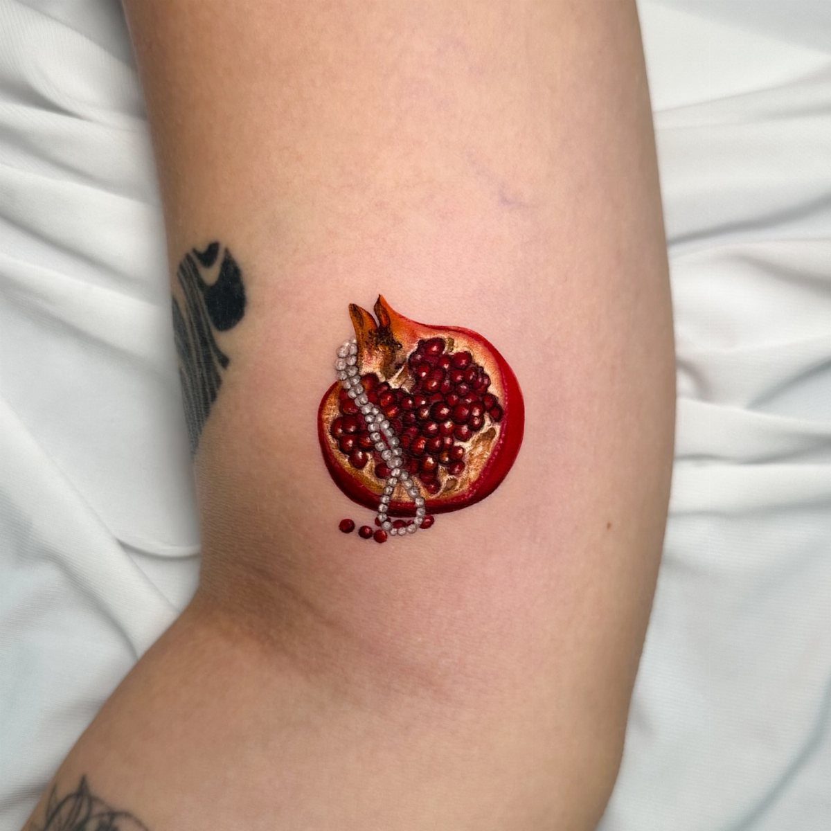 Pomegranate Tattoo Ideas: 11 Designs from Myth to Botanical 2 A realistic sliced open pomegranate tattoo with red seeds and white pearls on an arm.