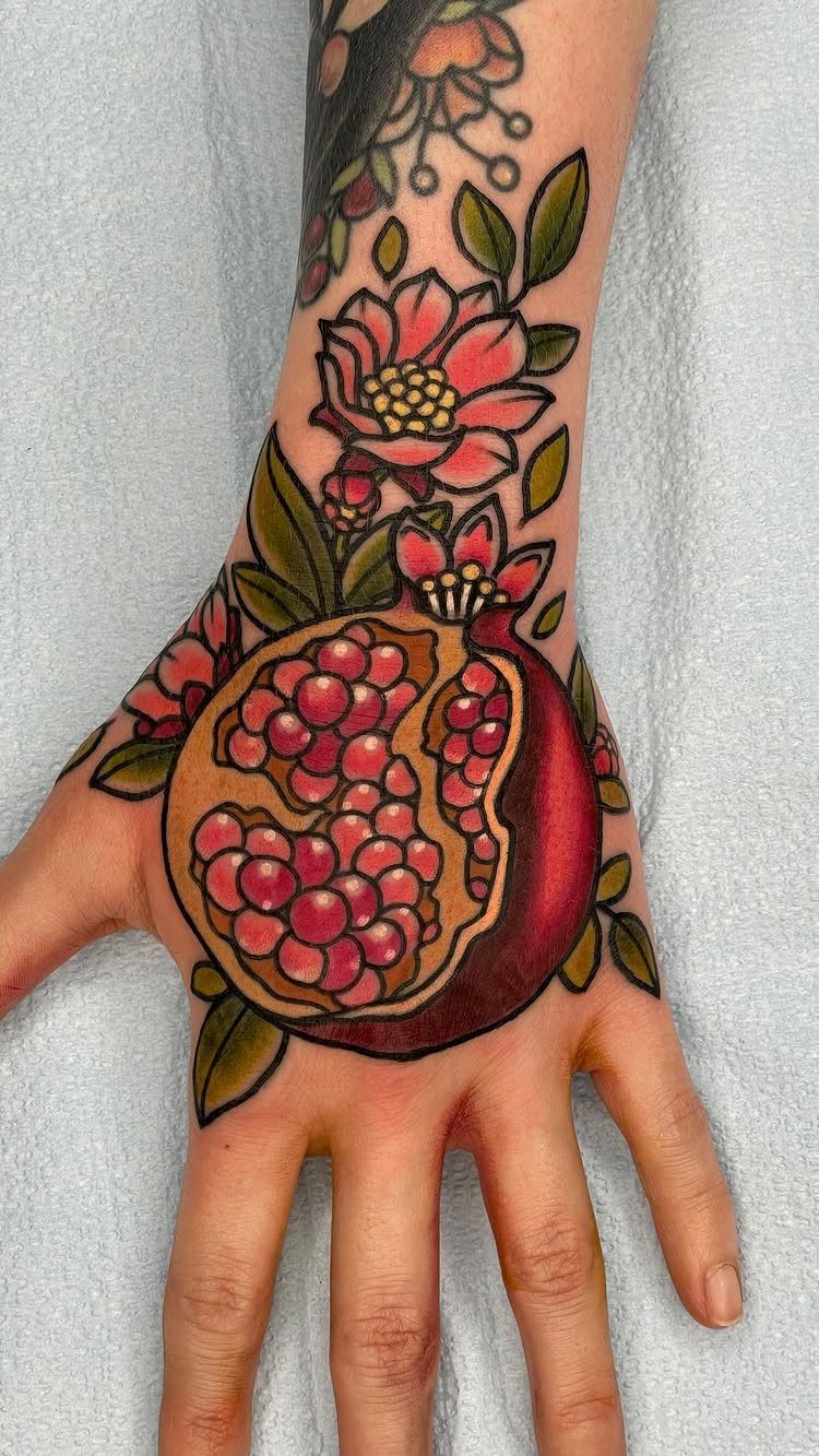 Pomegranate Tattoo Ideas: 11 Designs from Myth to Botanical 3 A traditional style pomegranate tattoo with red seeds and pink flowers on a person's hand.