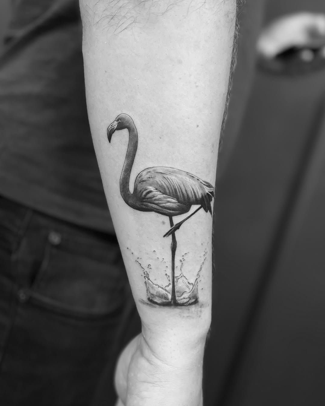 A realistic black and grey flamingo tattoo with water splashes on a person's forearm.