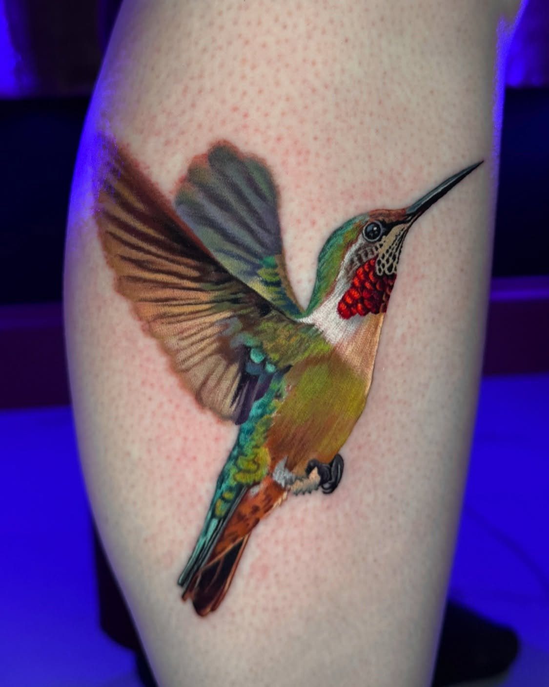 A detailed, colorful hummingbird tattoo with green and yellow feathers on human skin.
