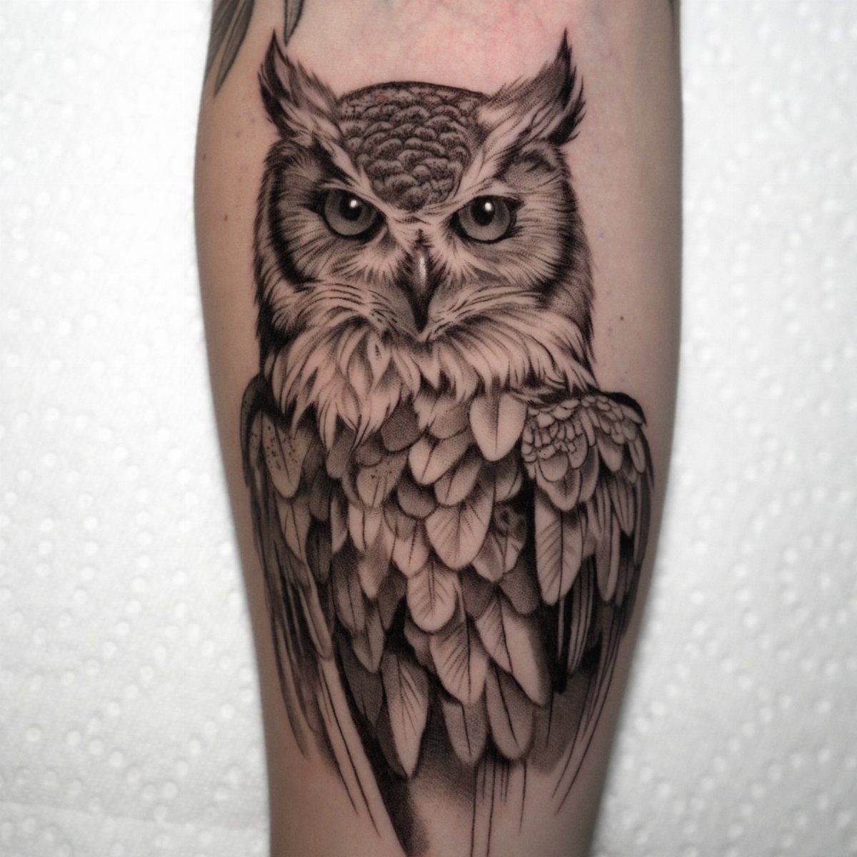 A realistic black and grey owl tattoo with detailed feathers on a person's skin.