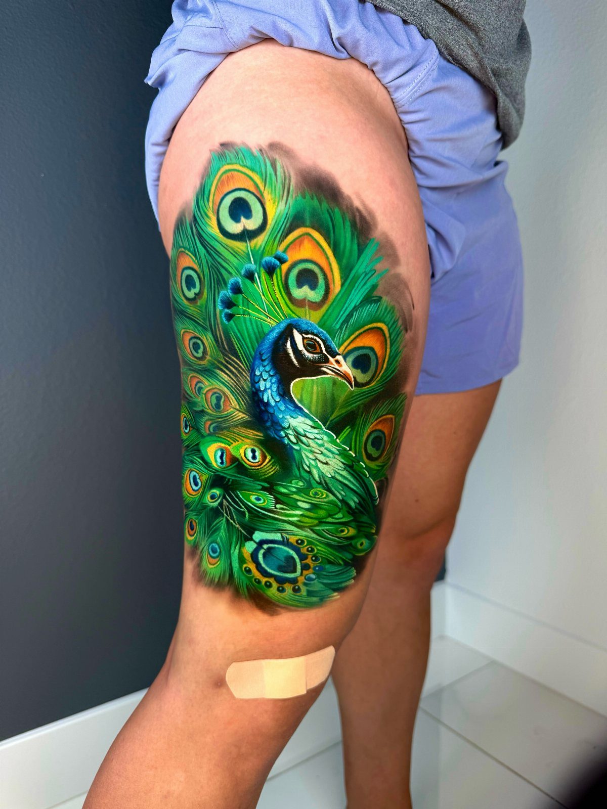 A colorful, realistic peacock tattoo with vibrant blue and green feathers on a person's thigh.