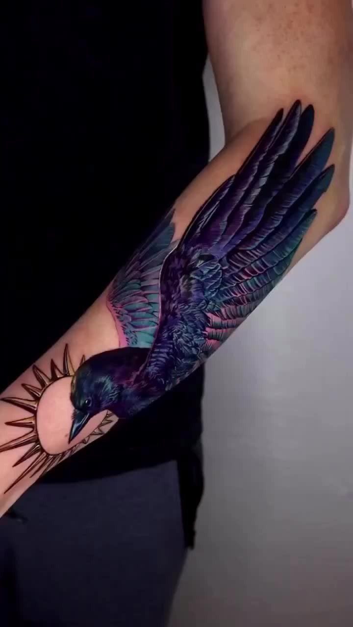 A detailed raven tattoo with iridescent purple and blue feathers on a person's forearm.