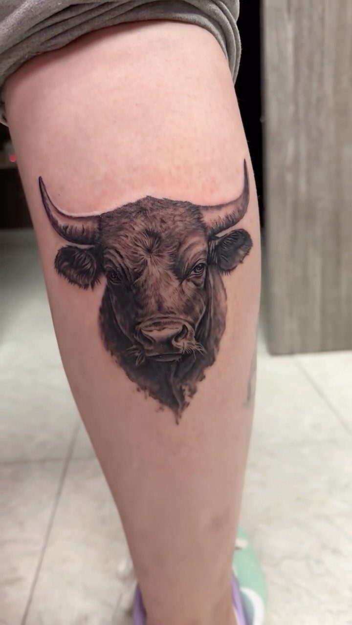 16 Realistic Bull Tattoo Ideas for Strength and Style 10 A detailed black and grey bull head tattoo on a person's leg.