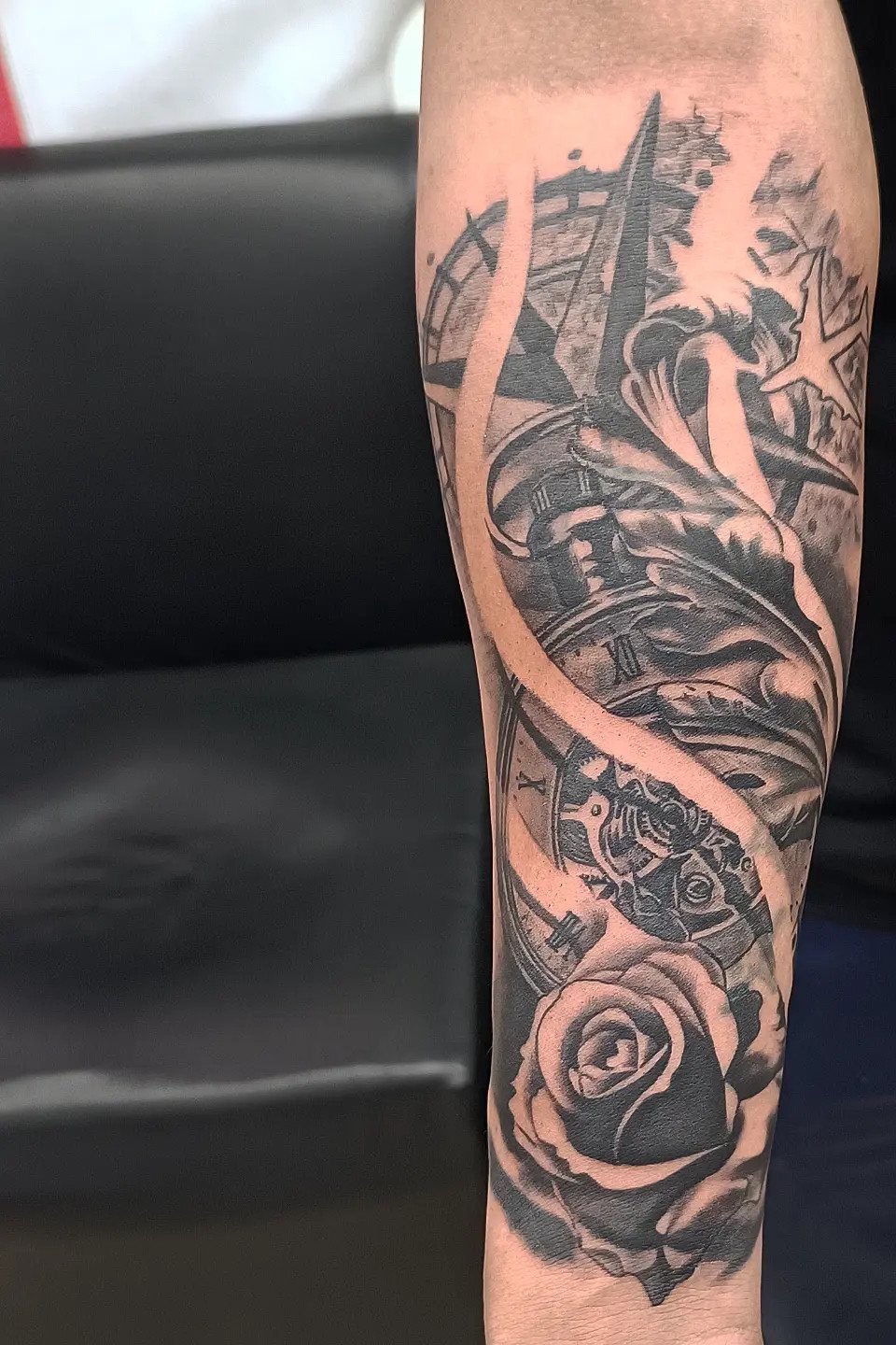 7 Rose with Pattern Tattoo Design Ideas for Ink That Stands Out 4 A black and grey tattoo on an arm featuring a rose, a compass, and flowing feather patterns.