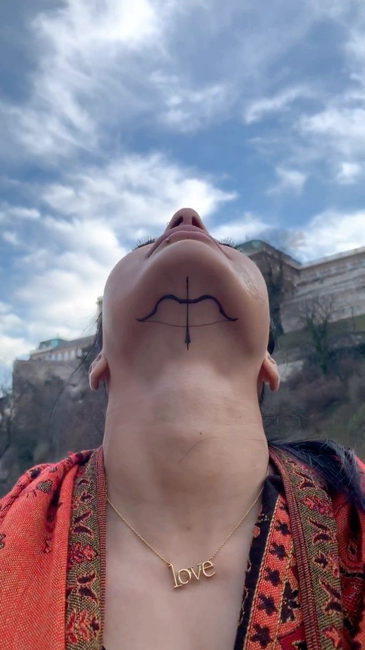 A person's chin featuring a small black bow tattoo against a cloudy blue sky.