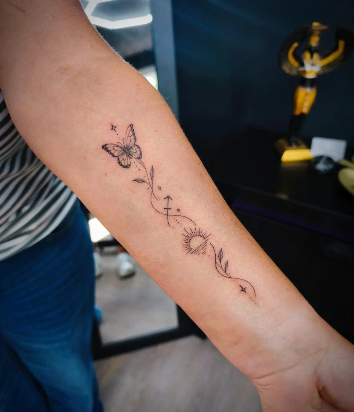 A fine line forearm tattoo featuring a butterfly, Sagittarius symbol, sun, and delicate leaves.
