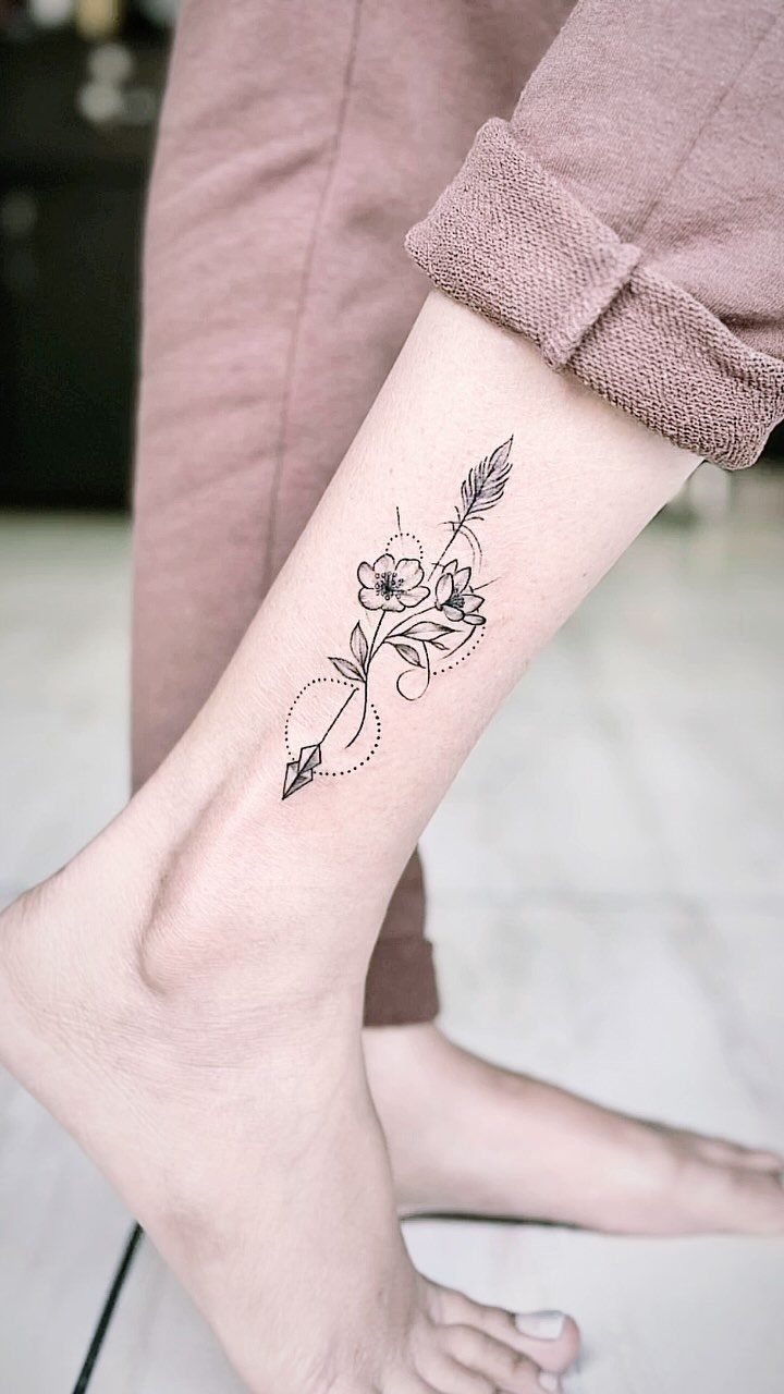 A fine line black ink arrow and flower tattoo on a person's lower leg.