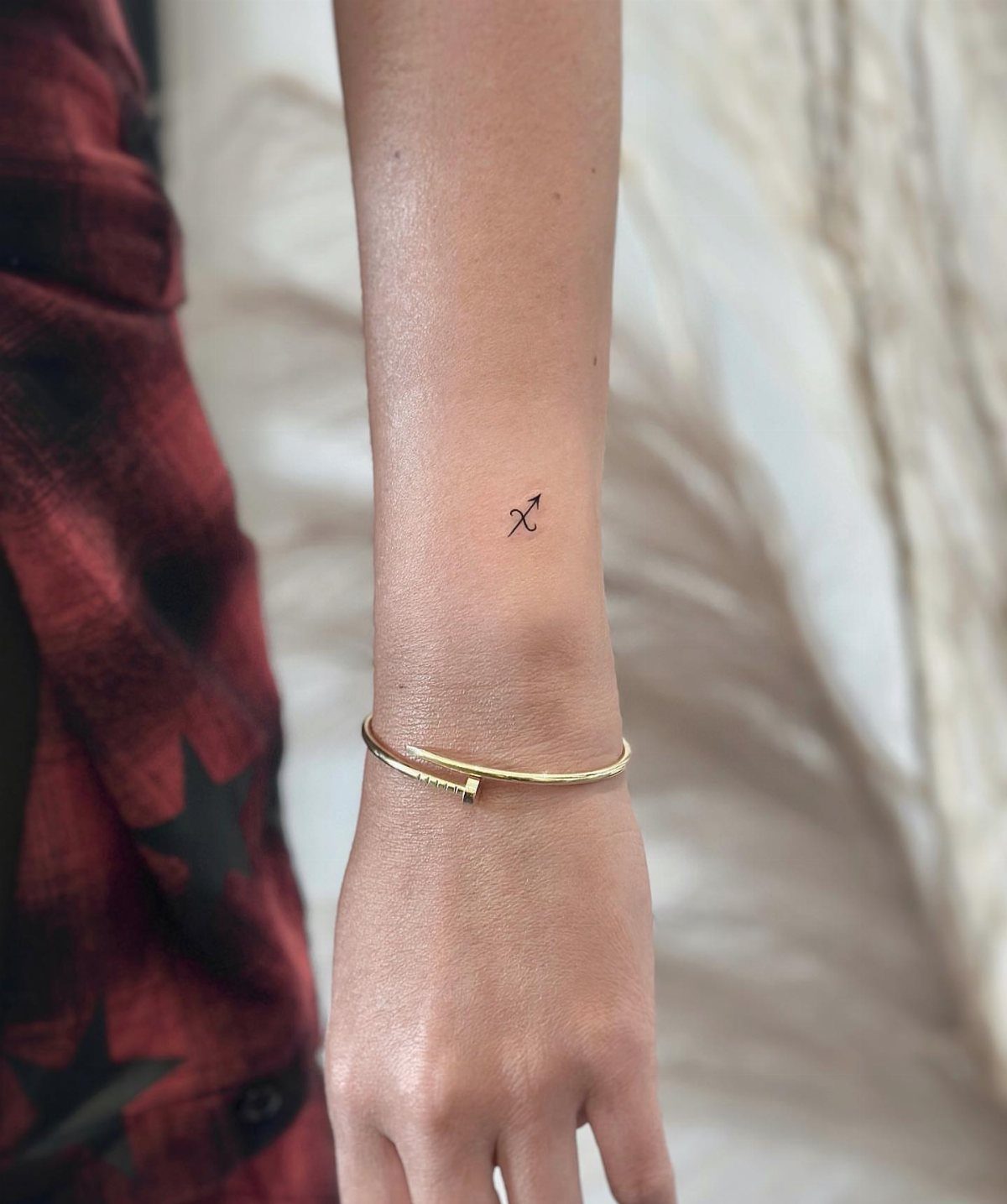 A small minimalist Sagittarius symbol tattoo on a person's inner wrist with a gold bracelet.