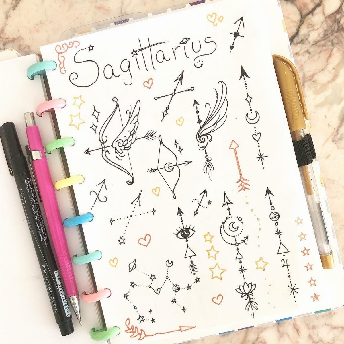 Hand-drawn Sagittarius tattoo sketches in a notebook with pens on a marble background.