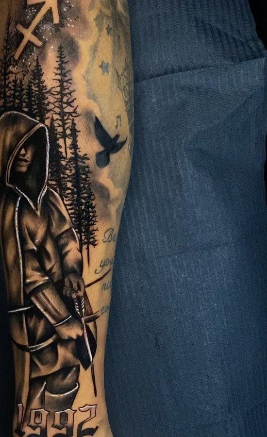A large black and grey tattoo on an arm depicting a hooded figure and forest scenery.