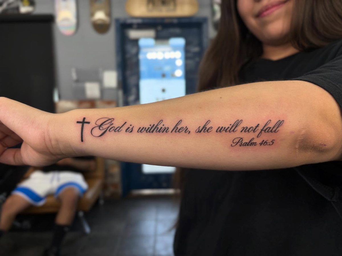 A forearm tattoo featuring a small cross and the biblical text from Psalm 46:5 in script.