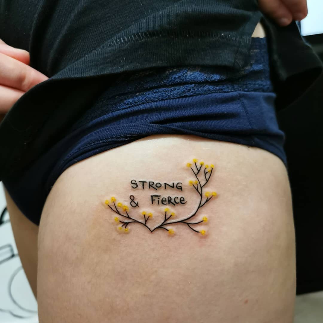 A small hip tattoo with the words strong and fierce surrounded by tiny yellow flowers.