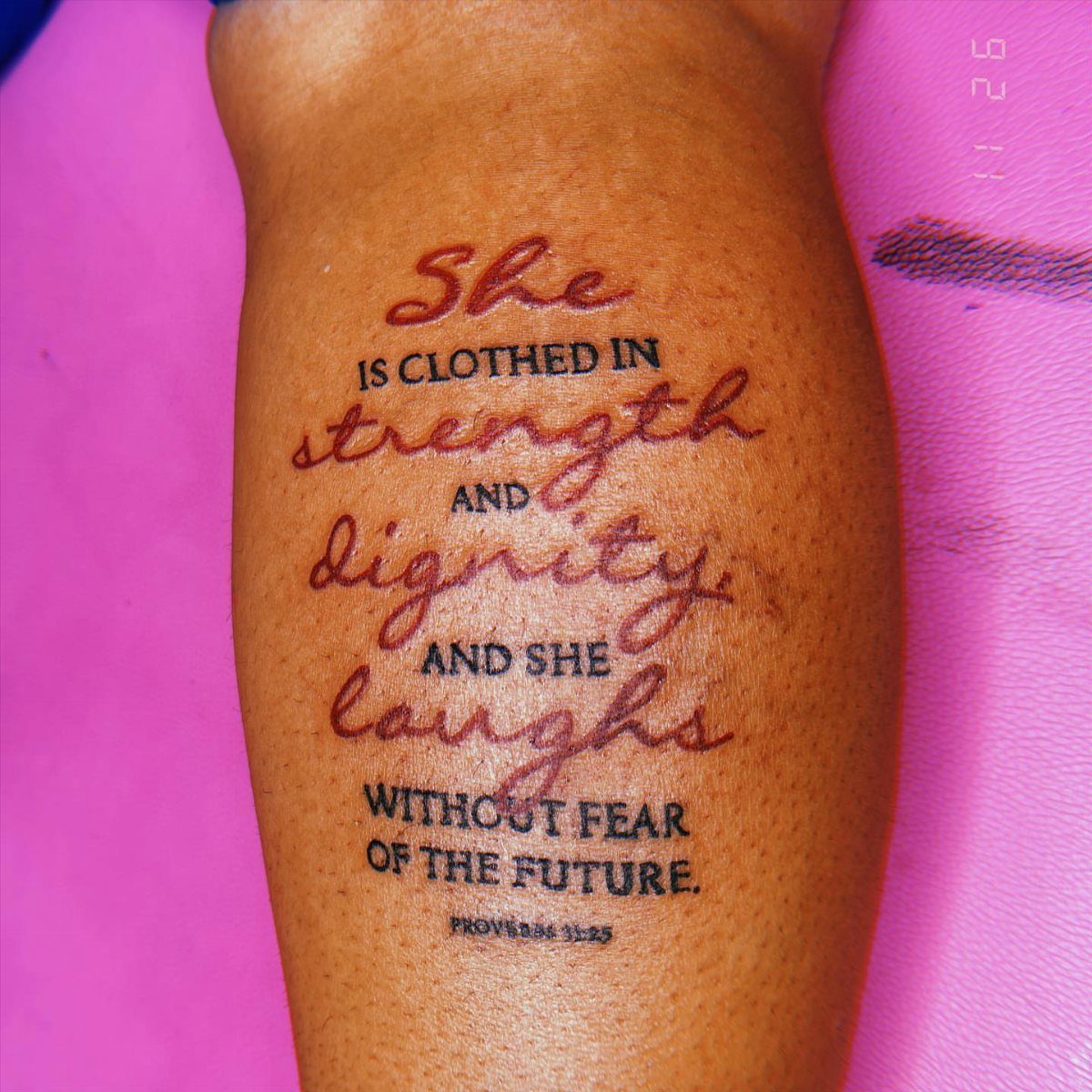 A script and serif text tattoo of Proverbs 31:25 on a person's skin.