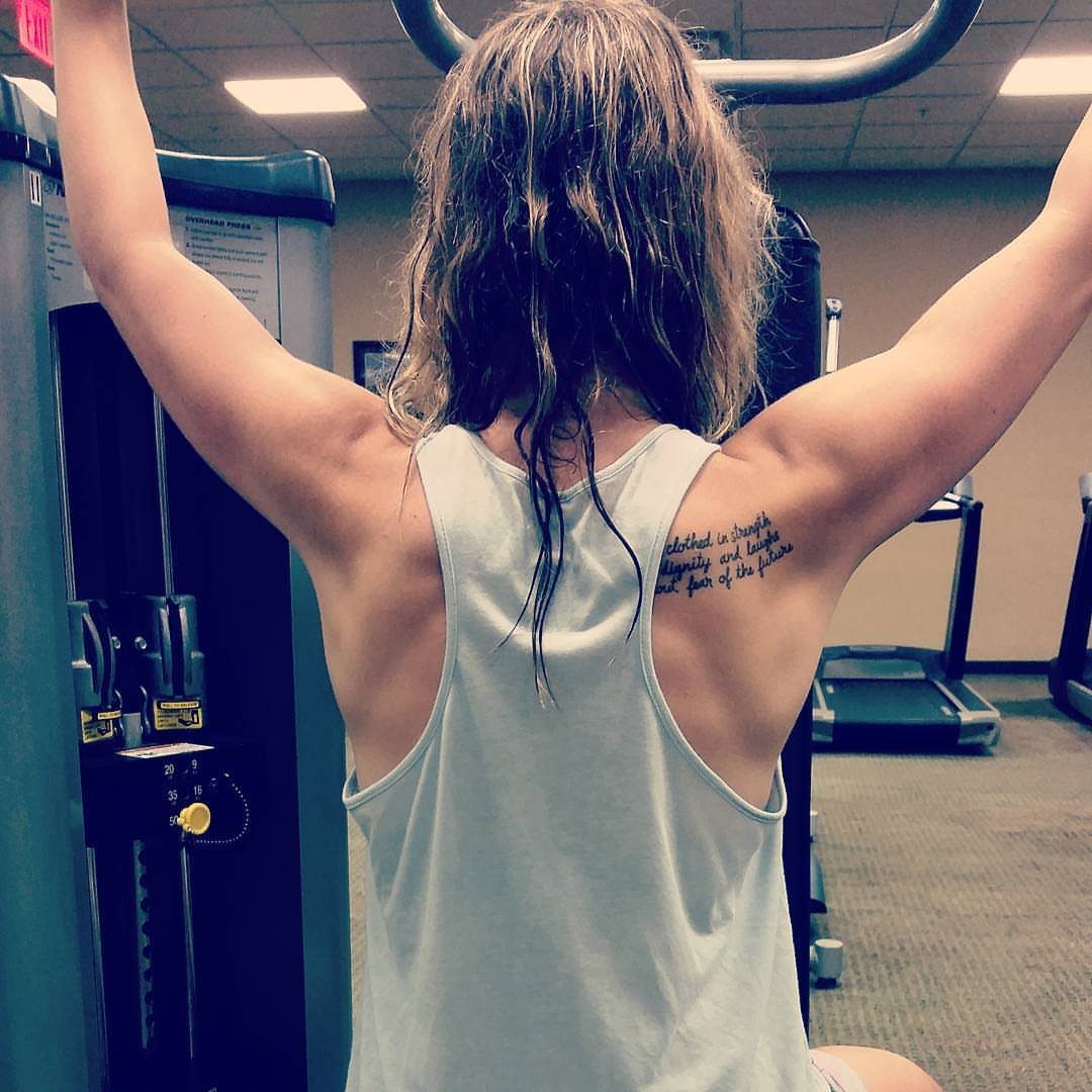 Woman in a white tank top showing a script tattoo on her right shoulder blade.
